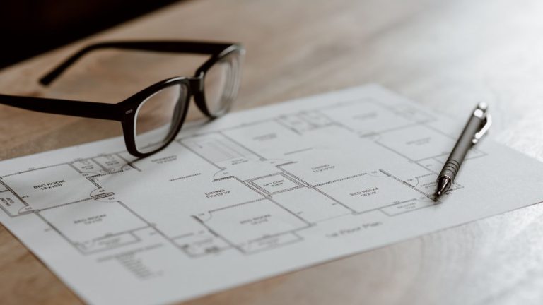 Eyeglasses and a pen resting on a floor plan on a wooden table in soft daylight